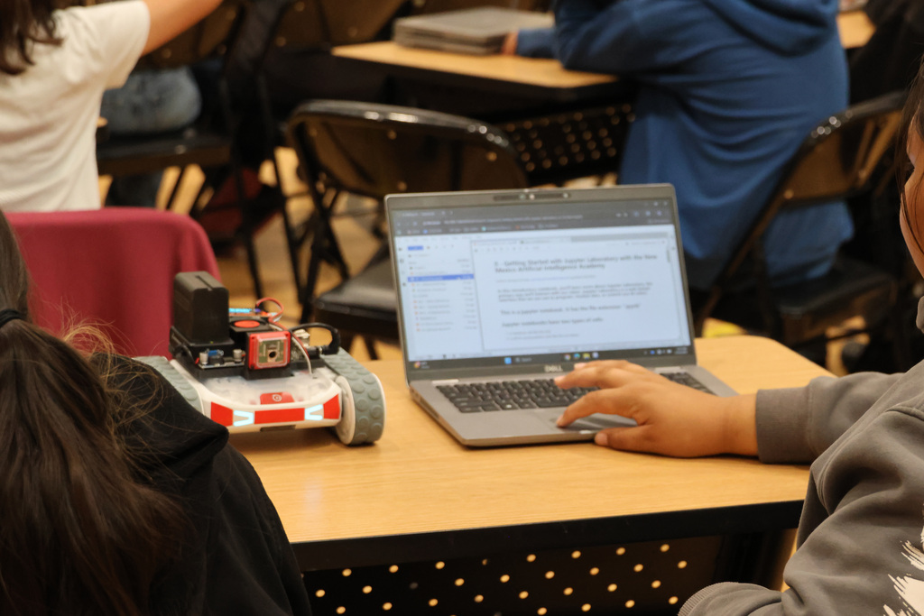 A student sits at a desk with a laptop and a small robot. A red chair is nearby.