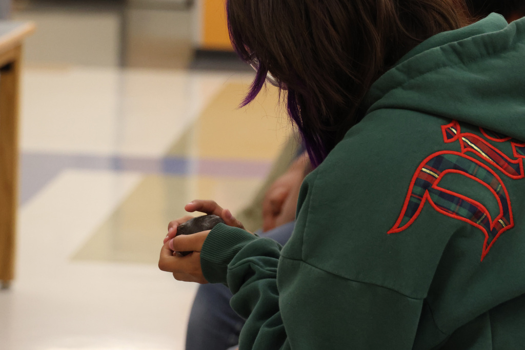 Person in green hoodie with red pattern on back, holding a small object, seated in a hallway.
