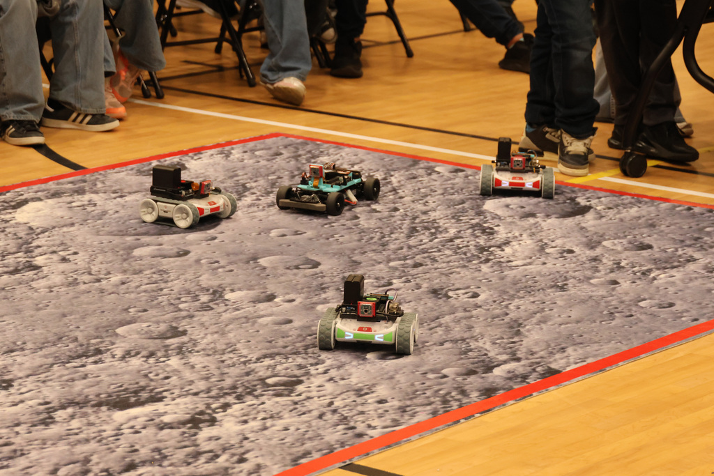 Small robots are on a floor marked with a moon's surface and red lines. People are standing around.