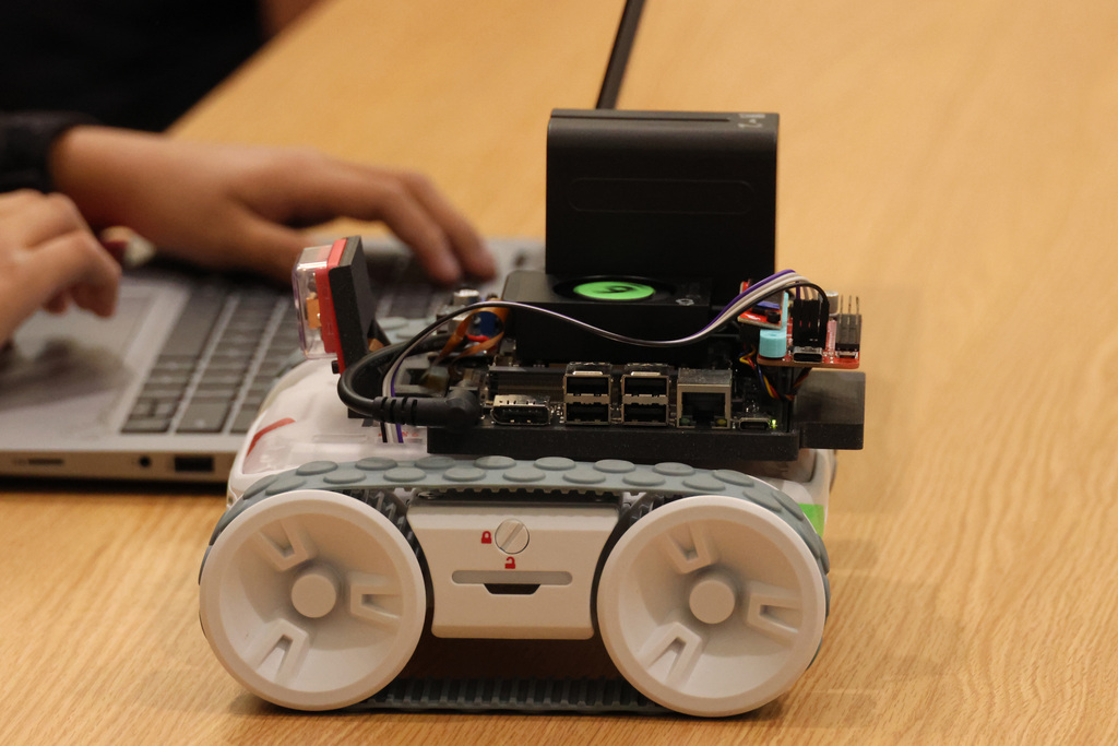 A person types on a laptop next to a robotic tank with wheels, positioned on a wooden table.