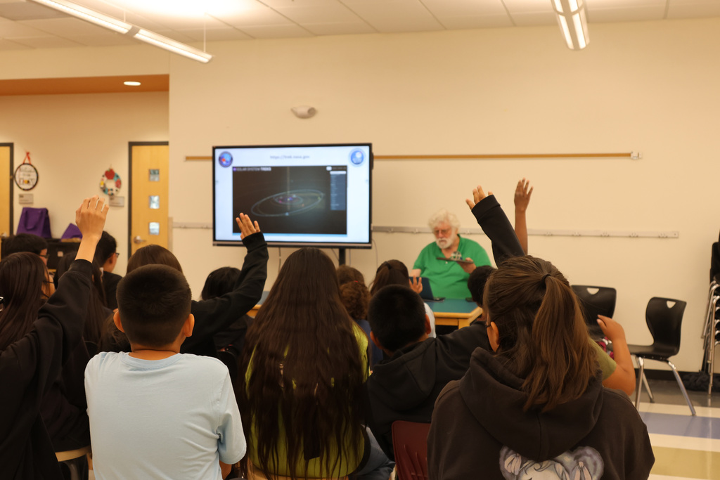 Classroom with projector; man in green shirt leads discussion. Students raise hands, seated in chairs.