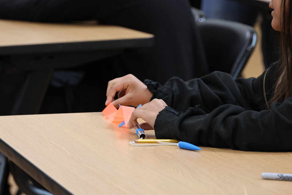 Person in a black long-sleeve shirt using orange paper and a blue marker on a desk.