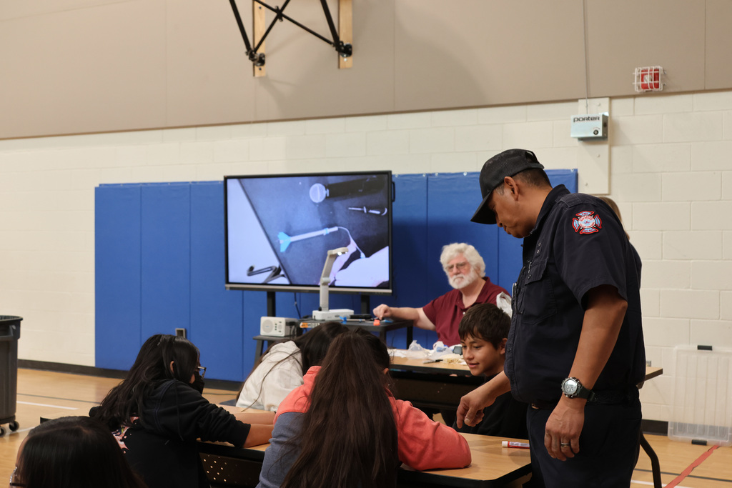 Fire safety training indoors with instructor and students. A large screen displays welding process. Participants seated around tables.