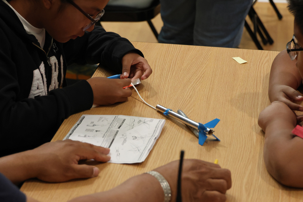 People work on a small plane model on a wooden table, one person in glasses is holding a wire.