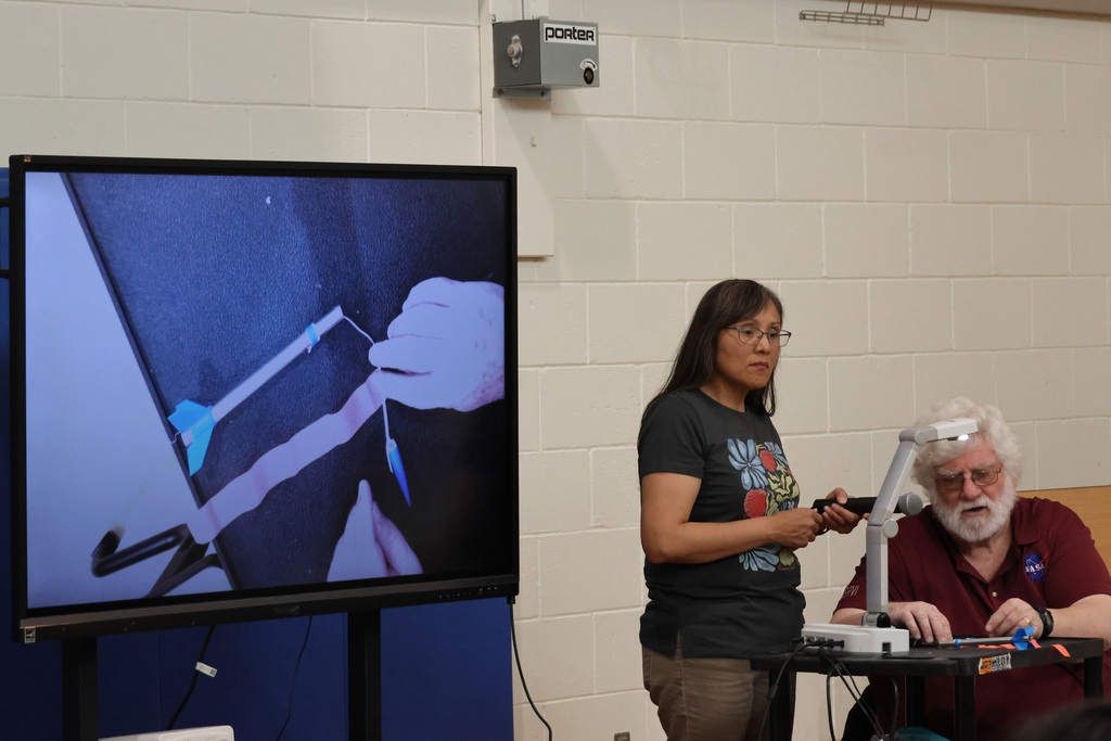 A woman speaks while standing in front of a screen showing a hand, with a man seated at a table.