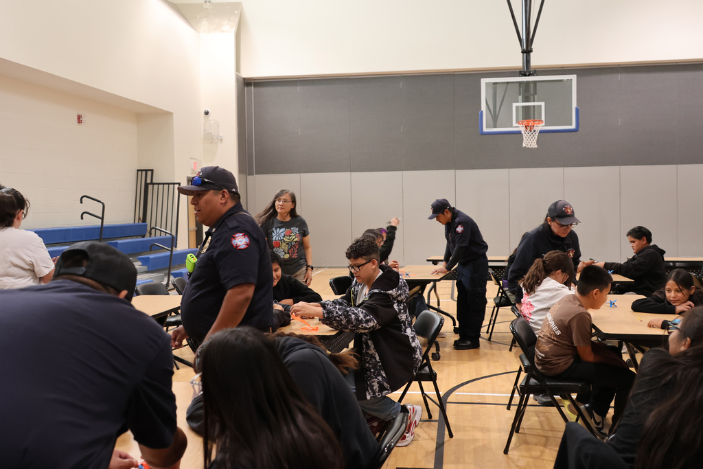 A group of people seated around tables in a gym, with adults standing. A basketball hoop is mounted on the wall.
