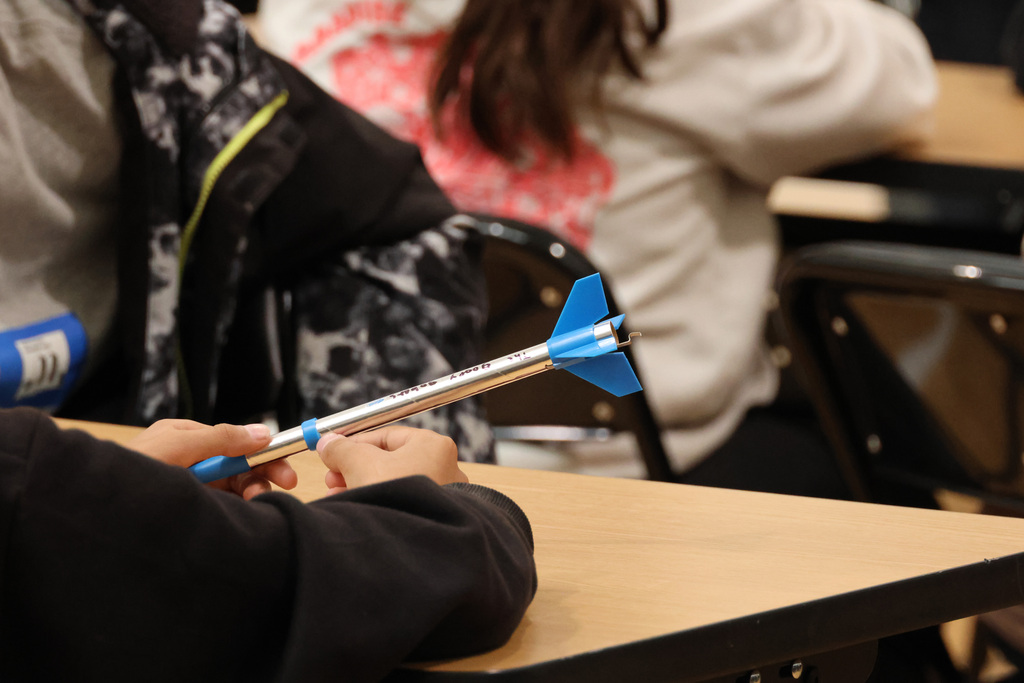A student in a black jacket holds a blue dart in a classroom, with another student in a white shirt seated nearby.