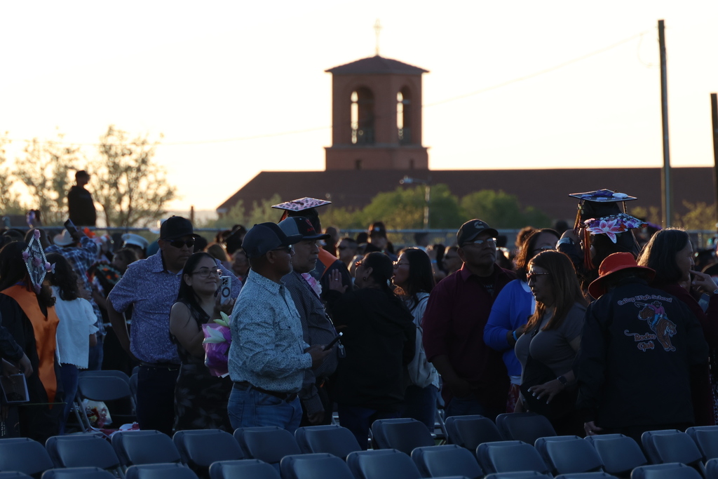 Crowd gathered in an outdoor amphitheater with church in the background. People wear hats, sunglasses, and some hold beverages.
