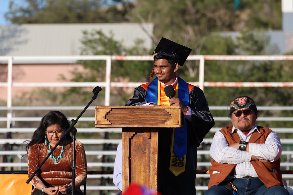 A man in a graduation gown speaks into a microphone at an outdoor podium. Two others watch from behind.