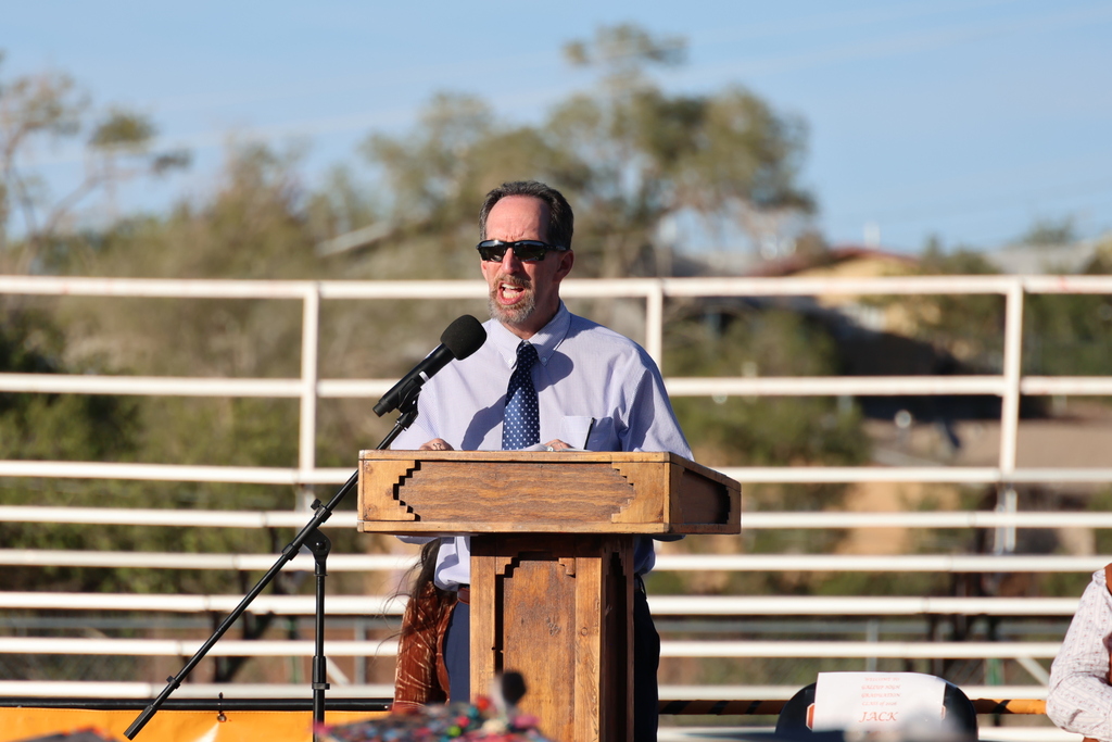 Man stands at podium with microphone, wearing sunglasses and a tie, with a white fence behind.