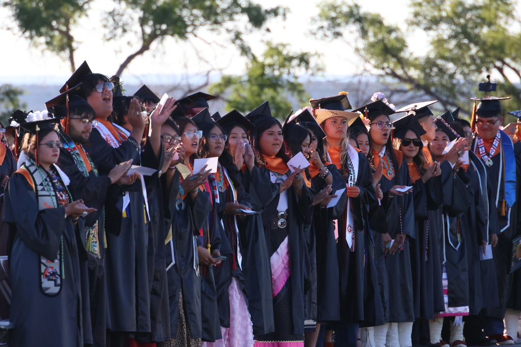 A line of graduates in academic regalia, some holding diplomas, stand outdoors with trees in the background.