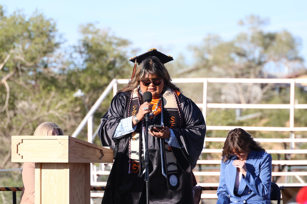 A person in a graduation gown speaks into a microphone. A woman sits behind them. Background shows bleachers.