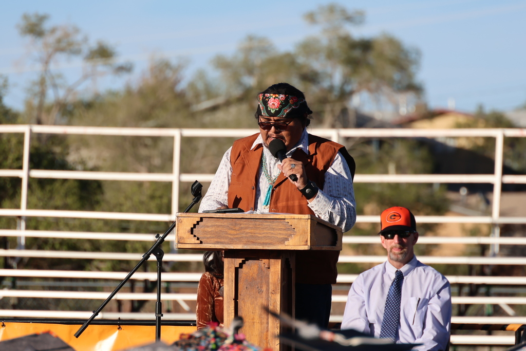Man at podium with microphone in hand, another man in long-sleeve shirt and tie standing behind.