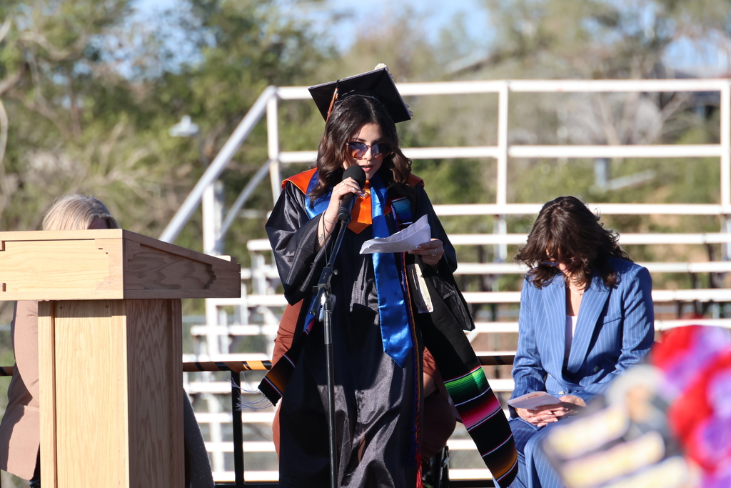 A woman with a graduation cap speaks into a microphone at a podium. People sit in bleachers.