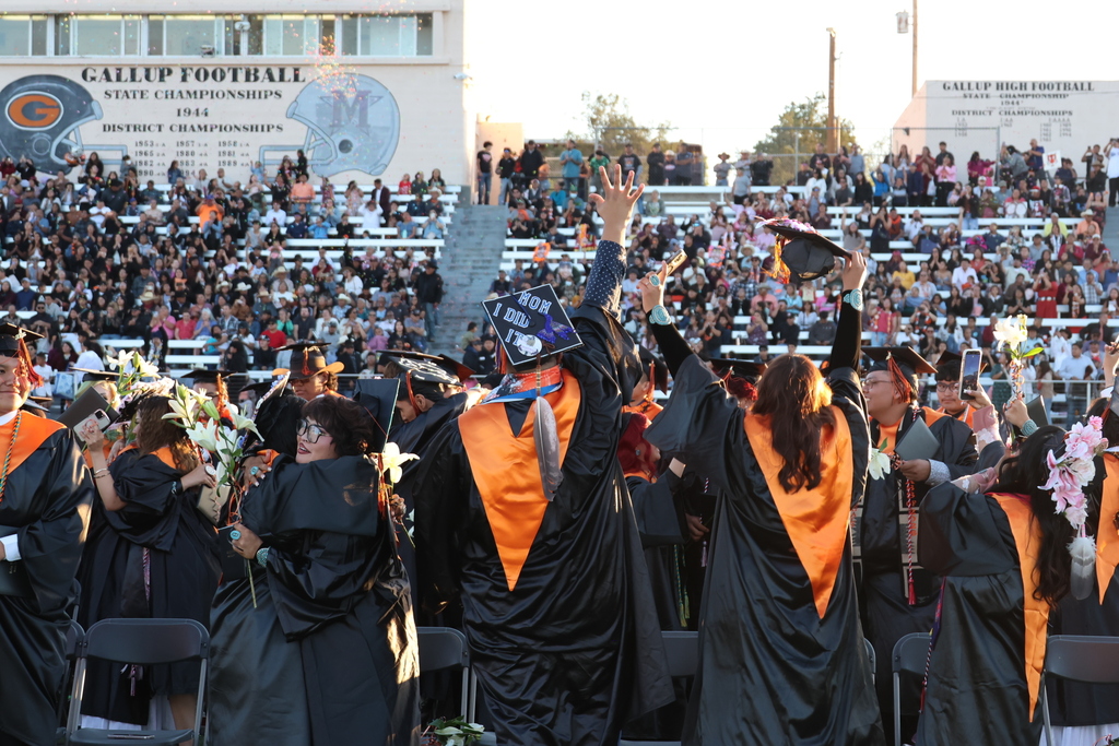 Graduates in black and orange gowns, some with hats, raising arms, and tossing hats in the air.
