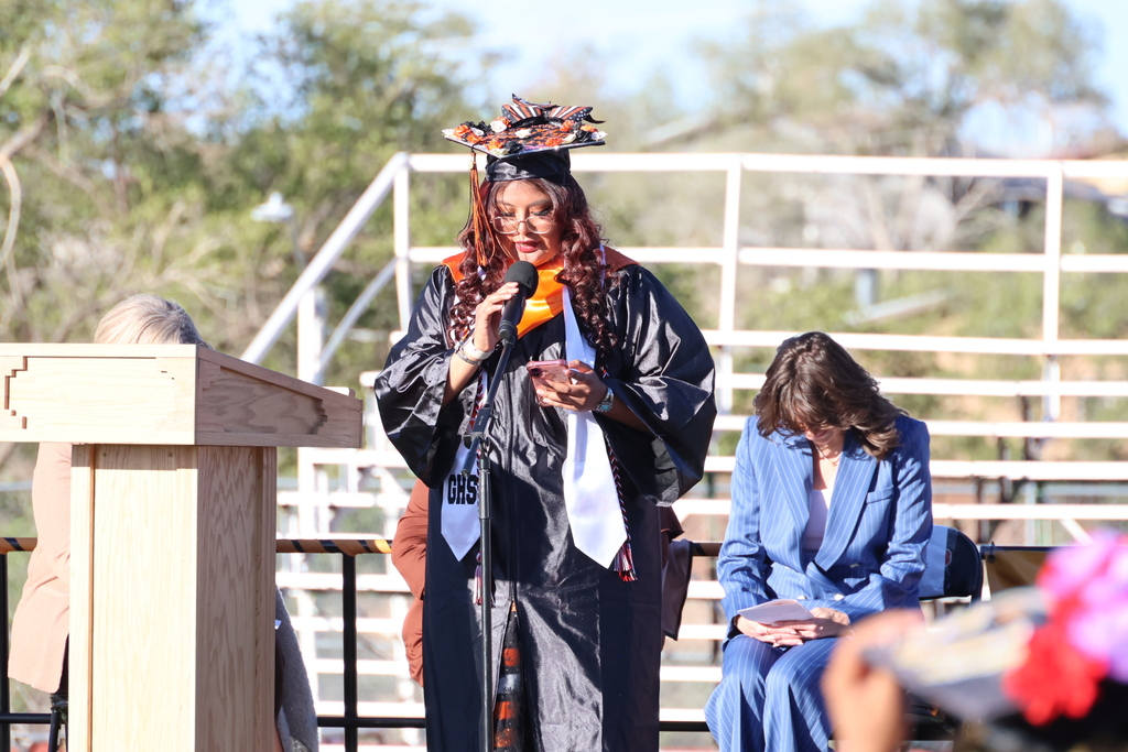 A woman in a cap and gown stands at a podium speaking into a microphone. Behind her, a seated woman in a blue blazer listens.