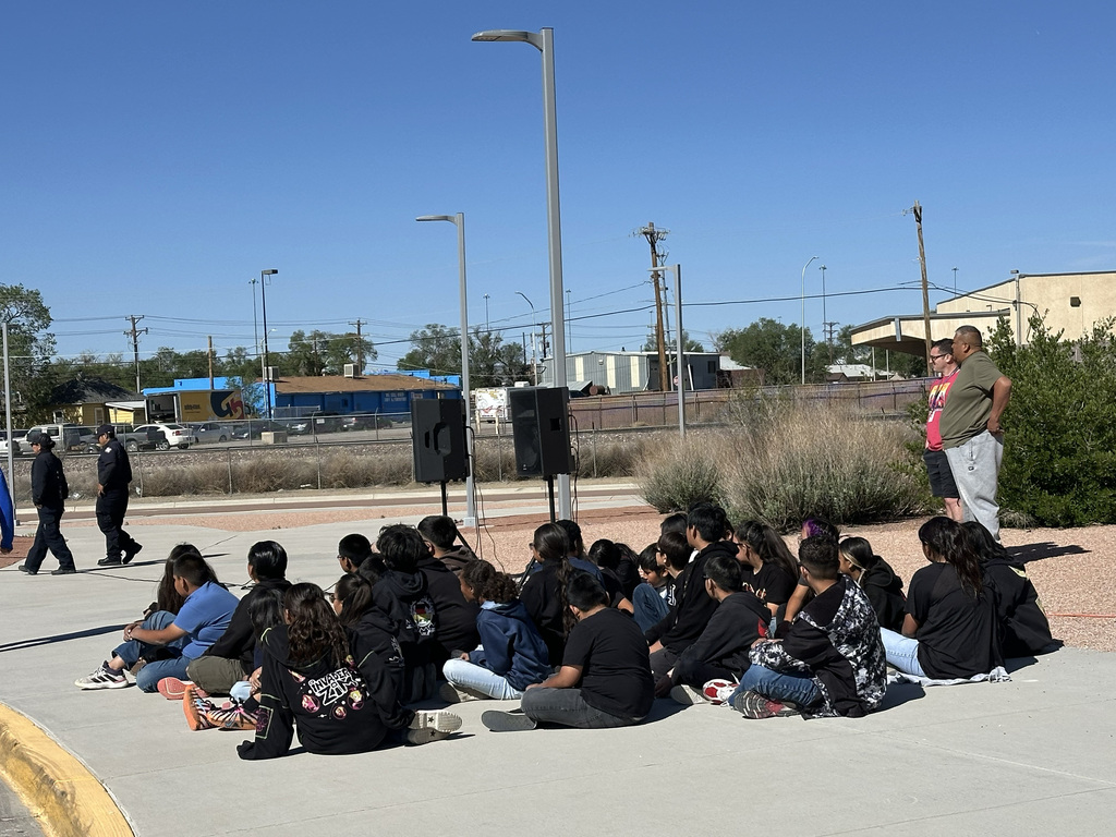 A group of people seated in a circle on concrete, with a speaker in the middle. Background includes a field, trees, and buildings.