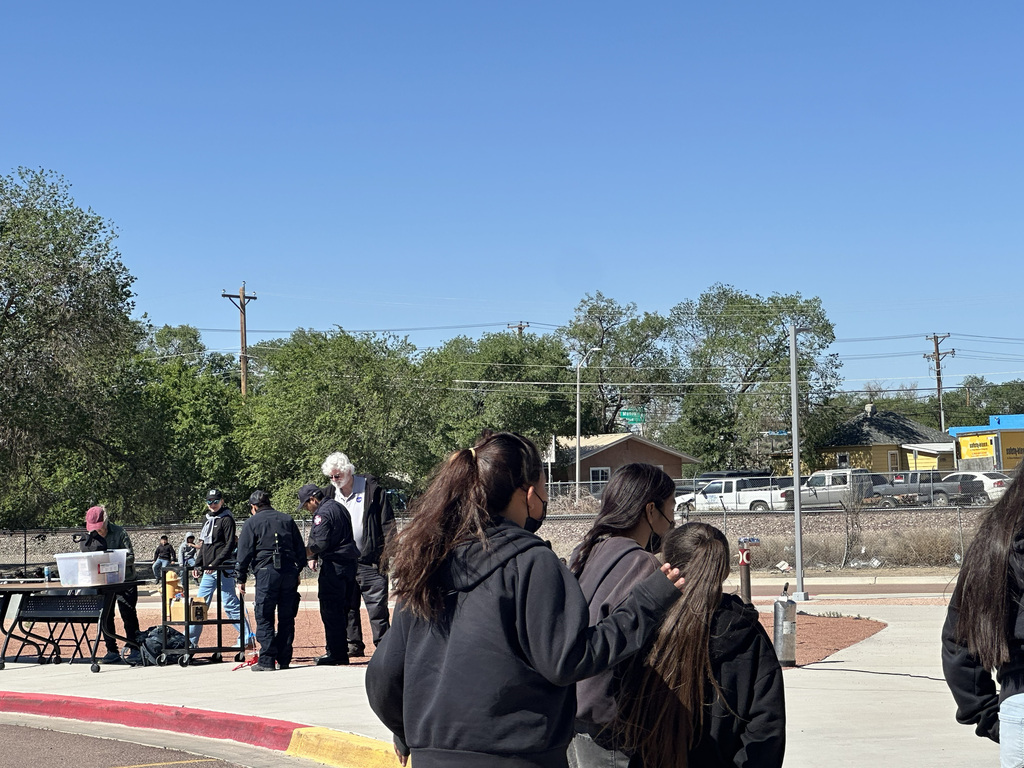 People gathered outdoors, with some standing near a table, others walking. Trees, buildings, and a clear sky in the background.