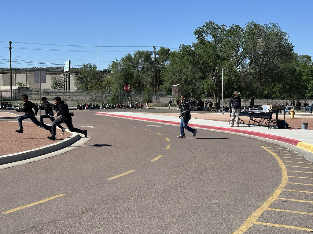 Several people jog on a paved road with yellow lines and a curb. Behind, a large building and trees are visible.