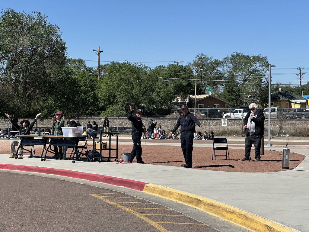People in uniforms stand in an outdoor area with a table, chairs, and trees in the background.