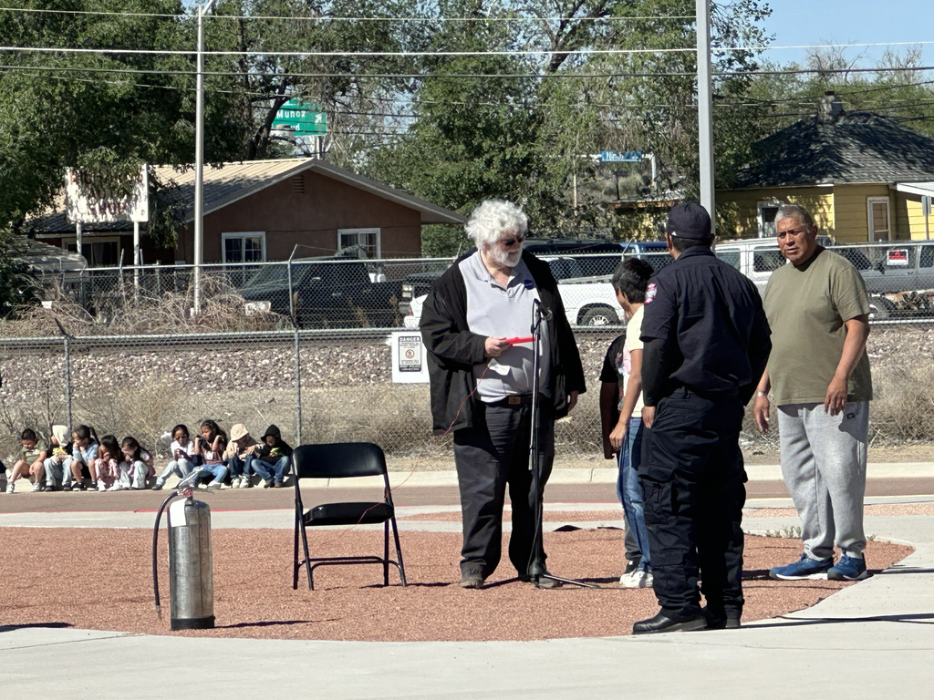 Four people stand in a circle, one holding a microphone. Behind them, a group of children sit on the ground. A fire extinguisher, chair, and fence are visible.