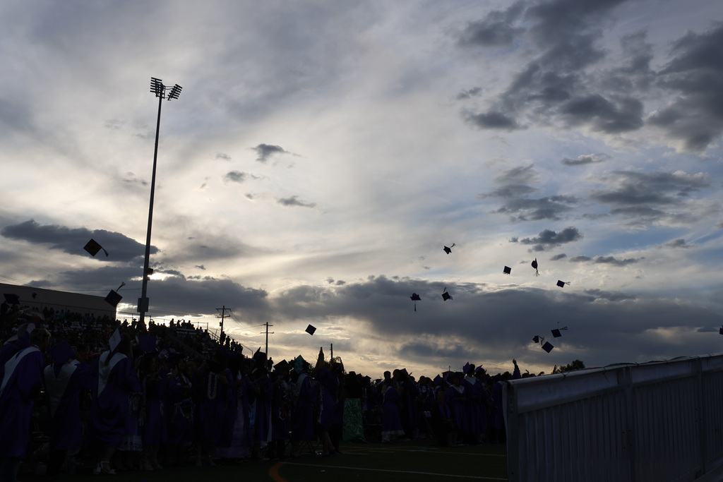 Graduates in blue gowns throw their caps into the air on a cloudy day. A white fence is visible in the foreground.