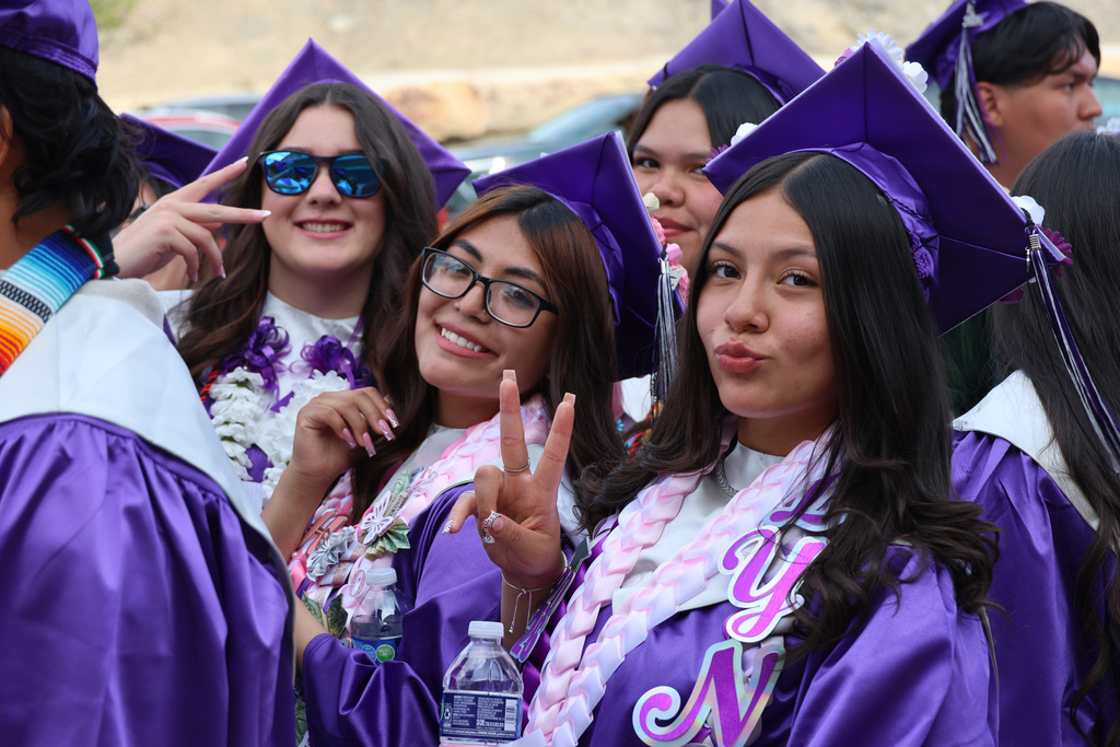 A group of young women in purple graduation gowns and caps smile and pose for a photo. Some wear glasses and make peace signs.