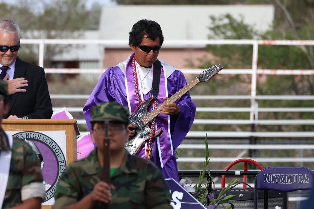A man in purple plays a guitar during a ceremony, accompanied by a military guard and a man at a podium.