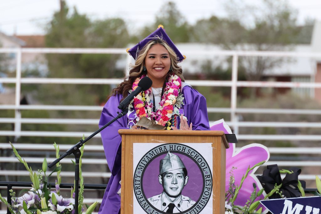 A woman in a purple gown stands at a podium, speaking into a microphone. She wears a cap and lei.