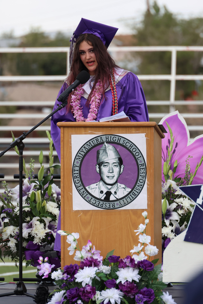 A woman wearing a purple graduation gown stands at a podium with flowers behind her, speaking into a microphone.