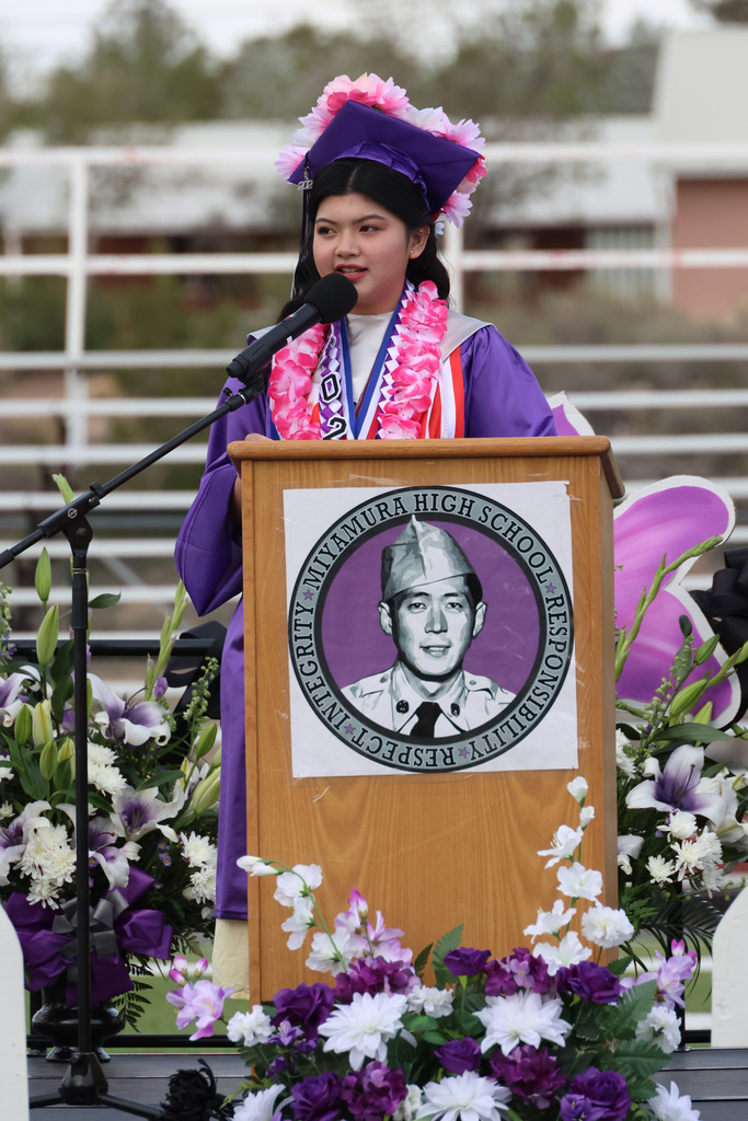 A woman in a graduation gown at a podium speaks into a microphone. A photo of a man is on the podium.