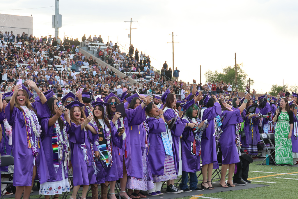 A large group of people in purple graduation gowns and caps on a stadium field, cheering.
