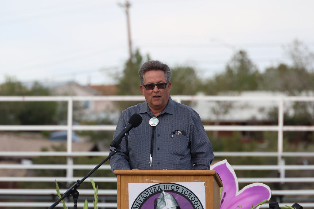 A man wearing sunglasses stands at a podium with a microphone, speaking. A logo is on the podium.