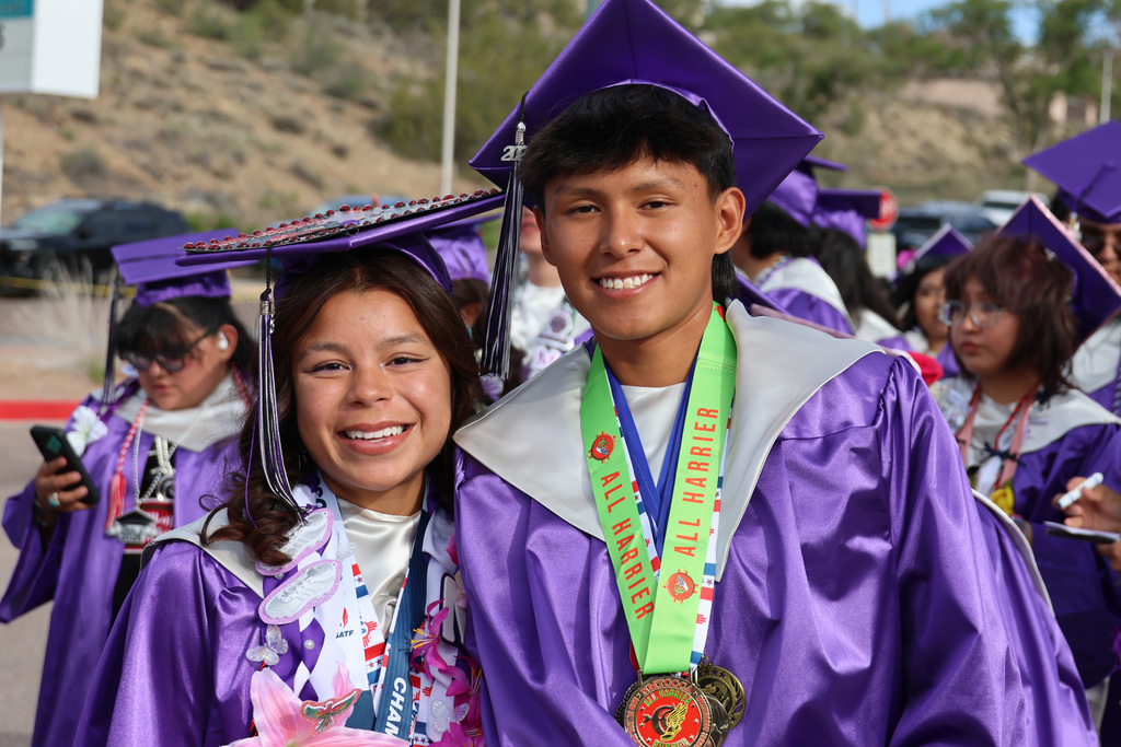 Graduates in purple gowns, one with a medal, smile for the camera. They stand in an outdoor area with a hill in the background.