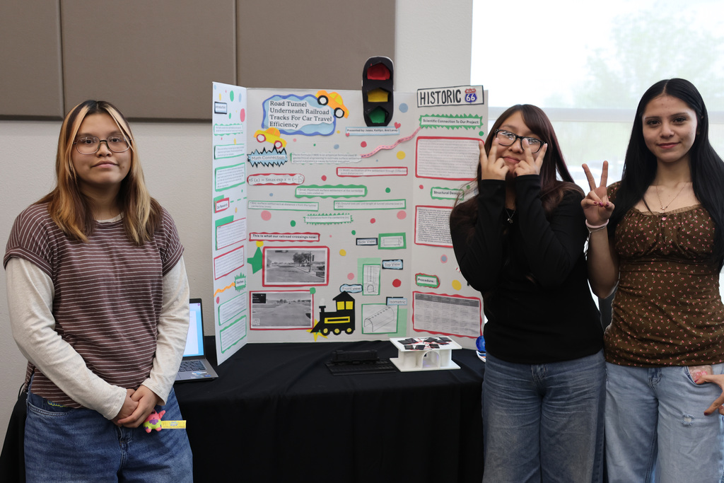 Three women stand next to a display board with a train map, buttons, and text. They smile and pose.