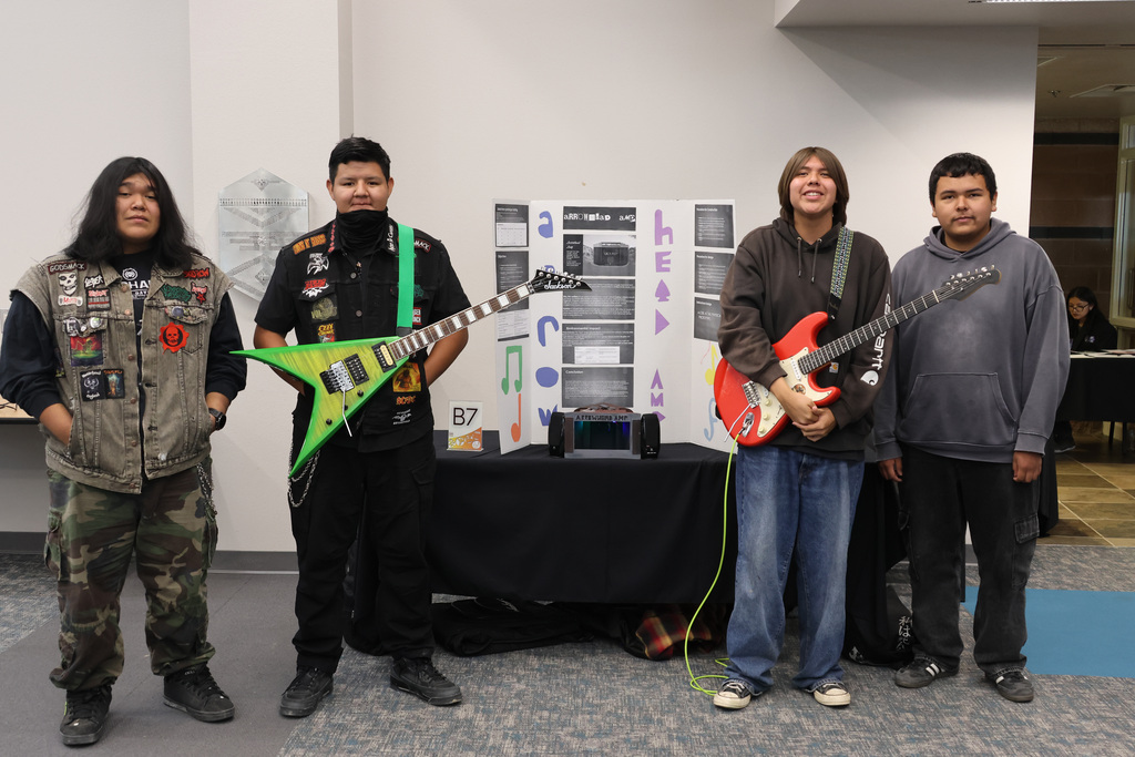 Four men stand in a room, each holding a guitar. The guitars are green, red, and black. Behind them, a black tablecloth covers a table.