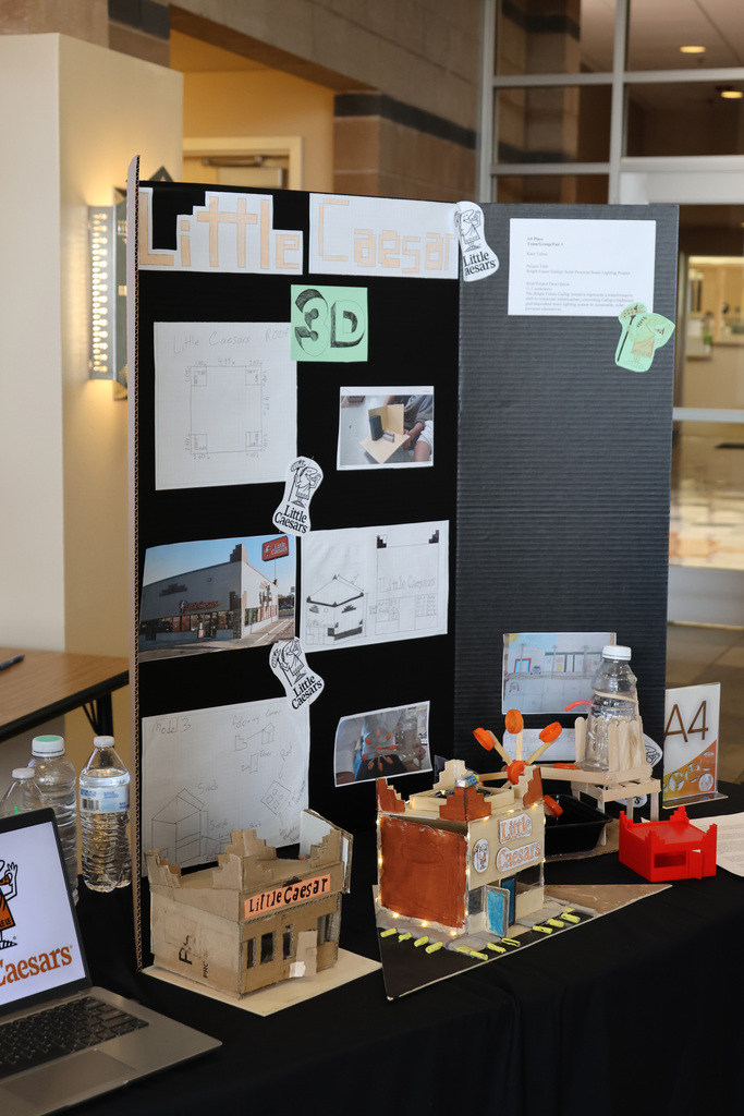 A table with a black tablecloth holds a laptop, cardboard models, and a display board with sketches and text.