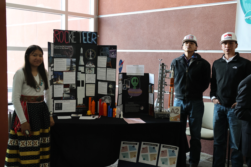 Three individuals standing by a table displaying a poster with "Rocketeer" and various rocket images and text.