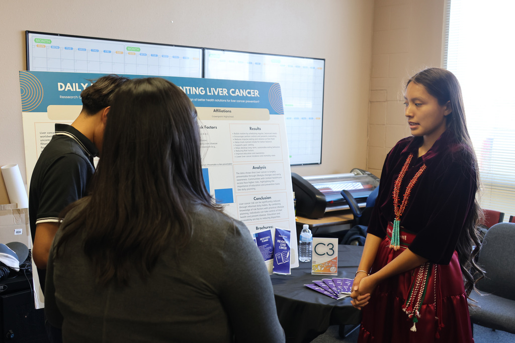 Three individuals stand in front of a poster on a table. The woman stands with hands clasped.