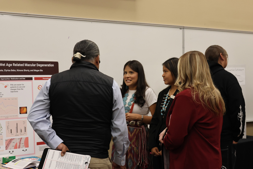 Six people view a poster on a white board, discussing. A person in a long skirt holds a folder.