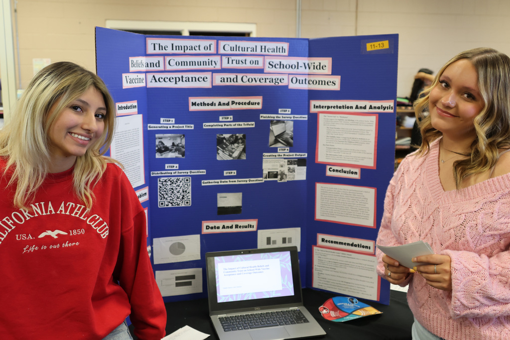Two women stand by a blue poster with text and images, a laptop open in front.