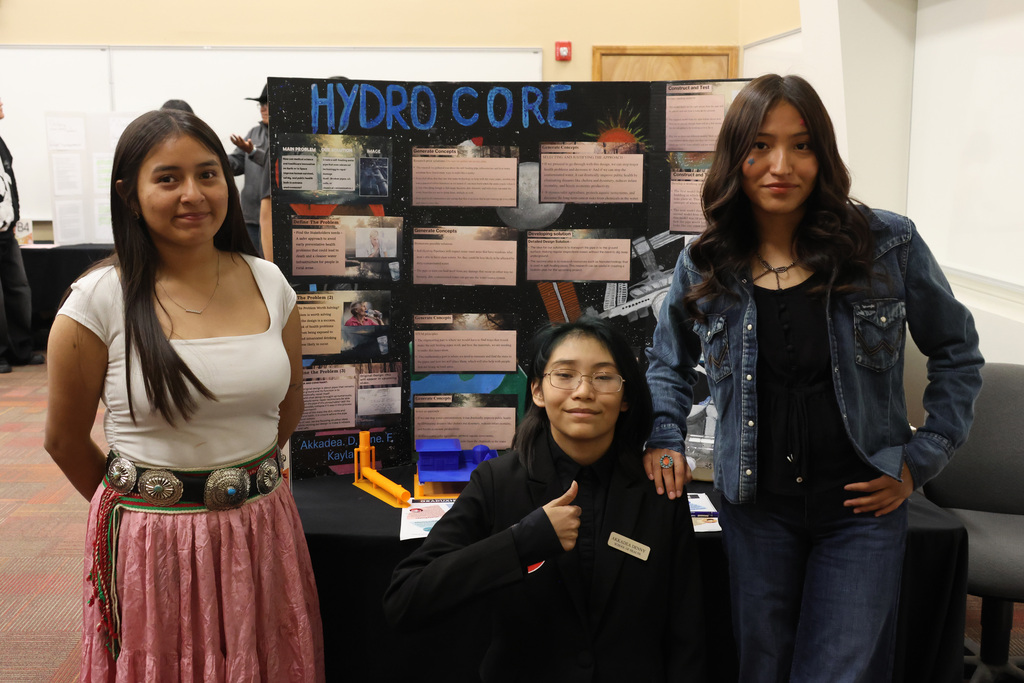Three women stand before a poster titled "Hydro Core" with a table behind them.