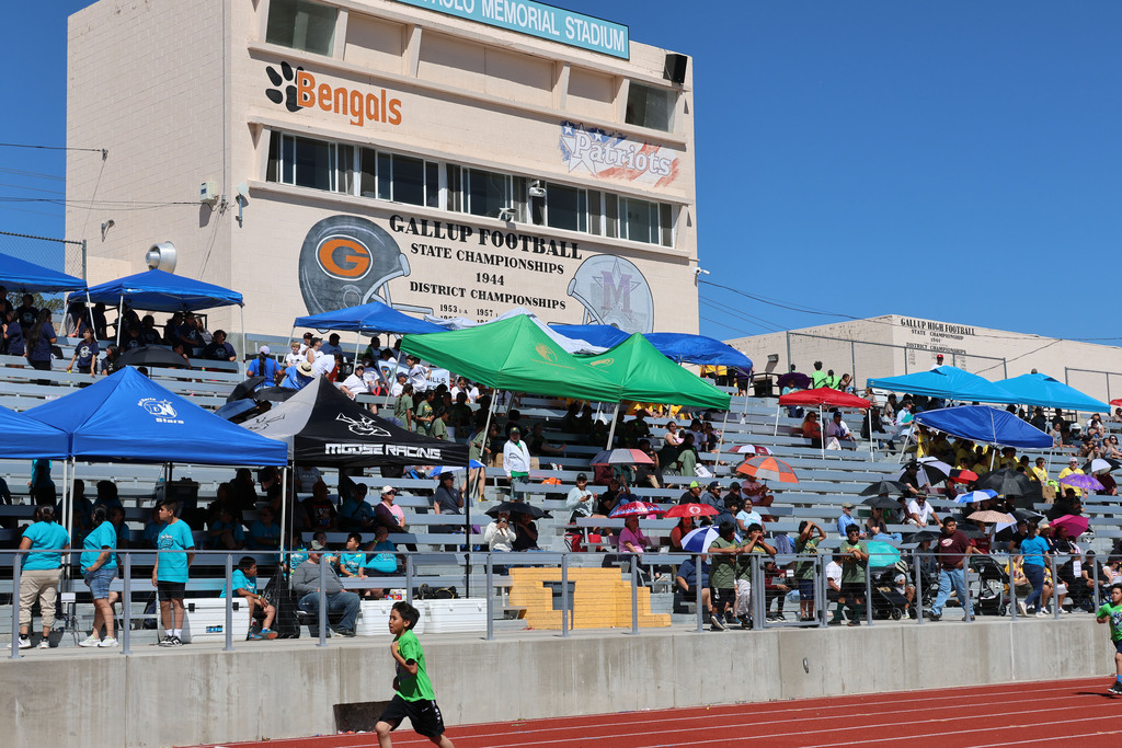 Spectators watch an outdoor track event from tiered seating. Large umbrellas of various colors provide shade.