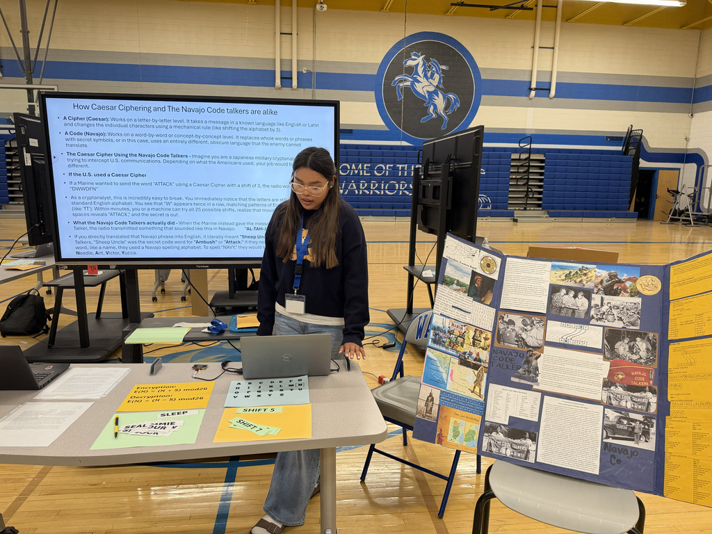 Person stands in gym, next to laptop and large screen, presenting a project. Display board with images and text nearby.