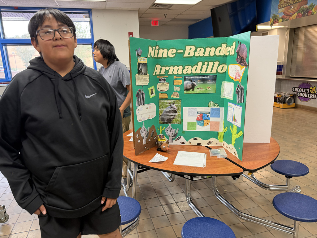 A person in a black jacket stands near a project board about nine-banded armadillos in a classroom.