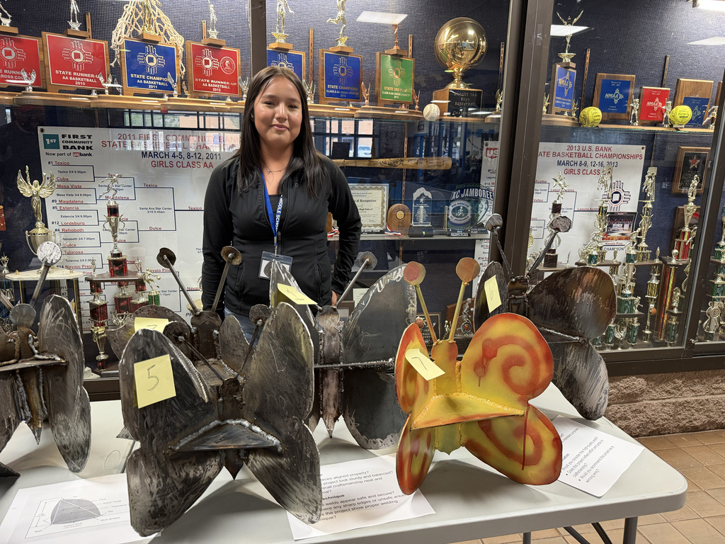 A woman stands behind a table of metal sculptures, with trophies and awards on display behind her.