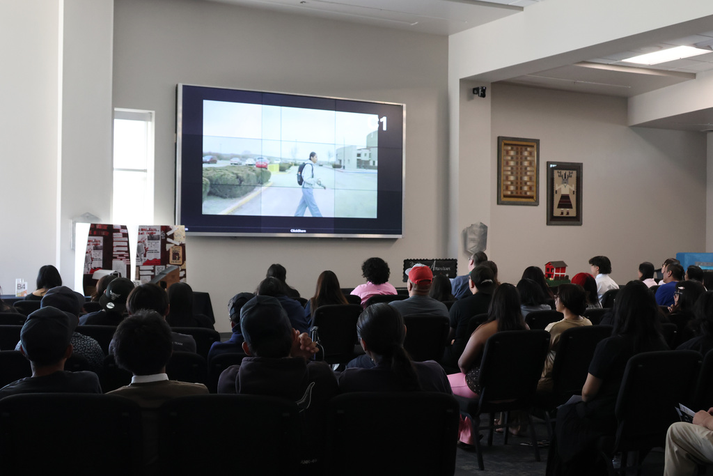 Room with a screen showing a person walking. An audience seated in chairs in front of the screen.