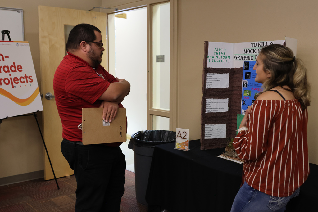 A man and a woman in a room with a table and display boards. The man holds a folder.