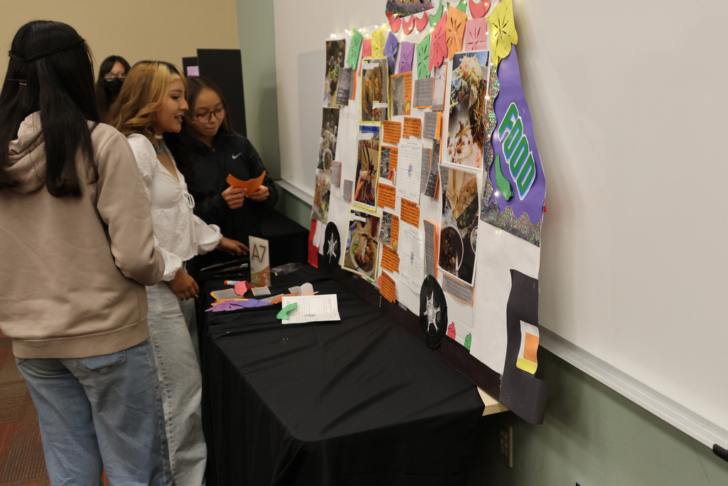 Three individuals gather around a table displaying a collage of images and text against a white wall.