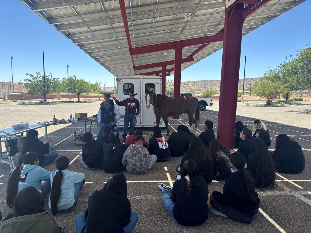 Group of individuals sit around under an awning. Two men stand with a horse. Trees and a building in the background.